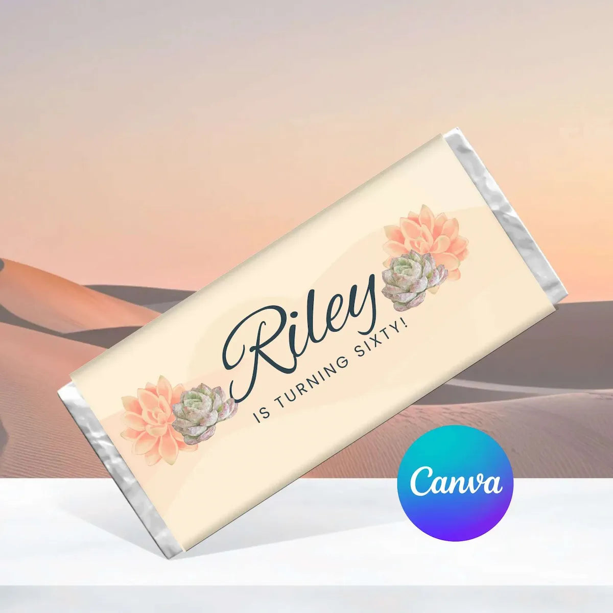 A custom wrapped chocolate bar with a cream wrapper featuring black script text reading 'Riley IS TURNING SIXTY!' and peach succulent illustrations, shown on a soft desert sunset background with a blue-to-purple Canva logo badge in the lower right.