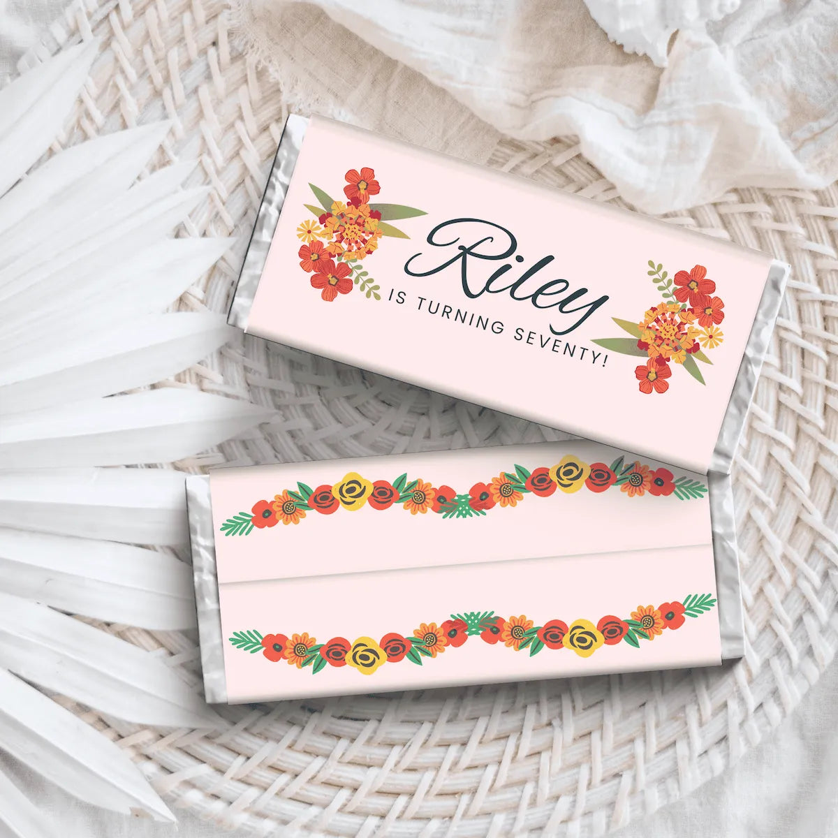 Two personalized chocolate bars with a light pink wrapper, decorated with orange and yellow floral designs and green leaves,  with Riley is Turning seventy text resting on a white woven placemat with white fabric in the background.