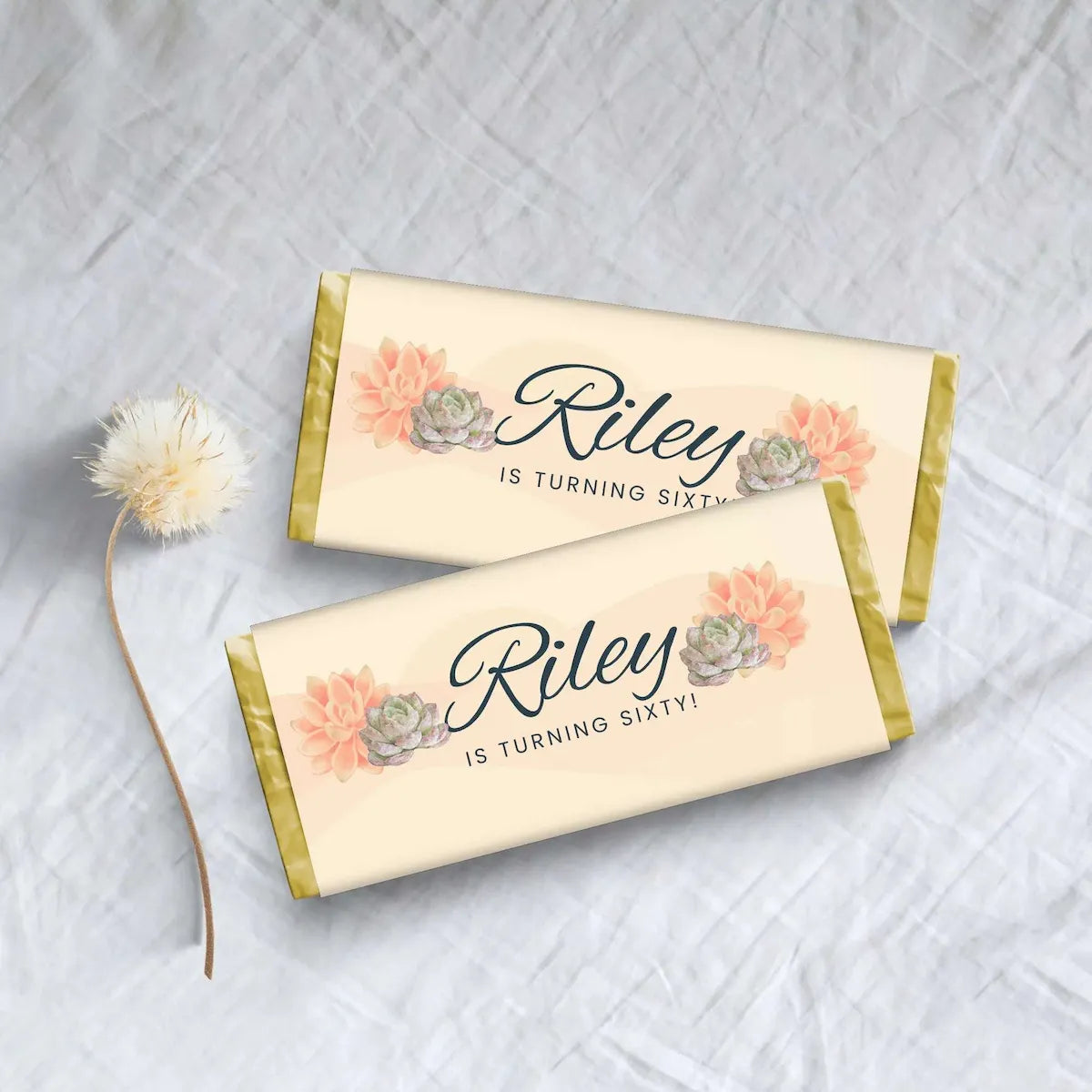 Two personalized candy bars wrapped in cream paper with gold foil edges, decorated with peach succulents and the text 'Riley IS TURNING SIXTY!', arranged on a light fabric background with a small dandelion stem.