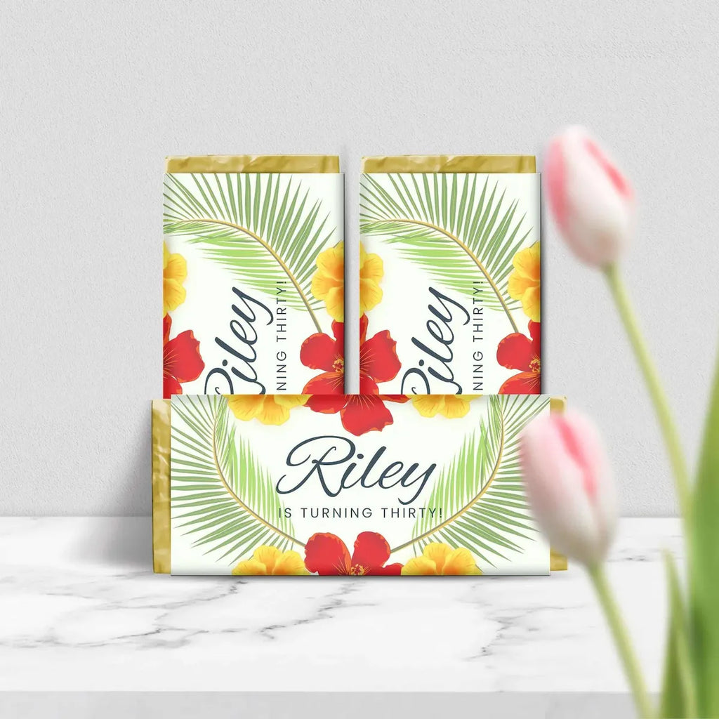 Three tropical-themed birthday cake toppers or wraps for a 30th birthday, featuring palm leaves, hibiscus flowers, and the name 'Riley' on a white background, styled on a marble surface with tulips in the foreground.