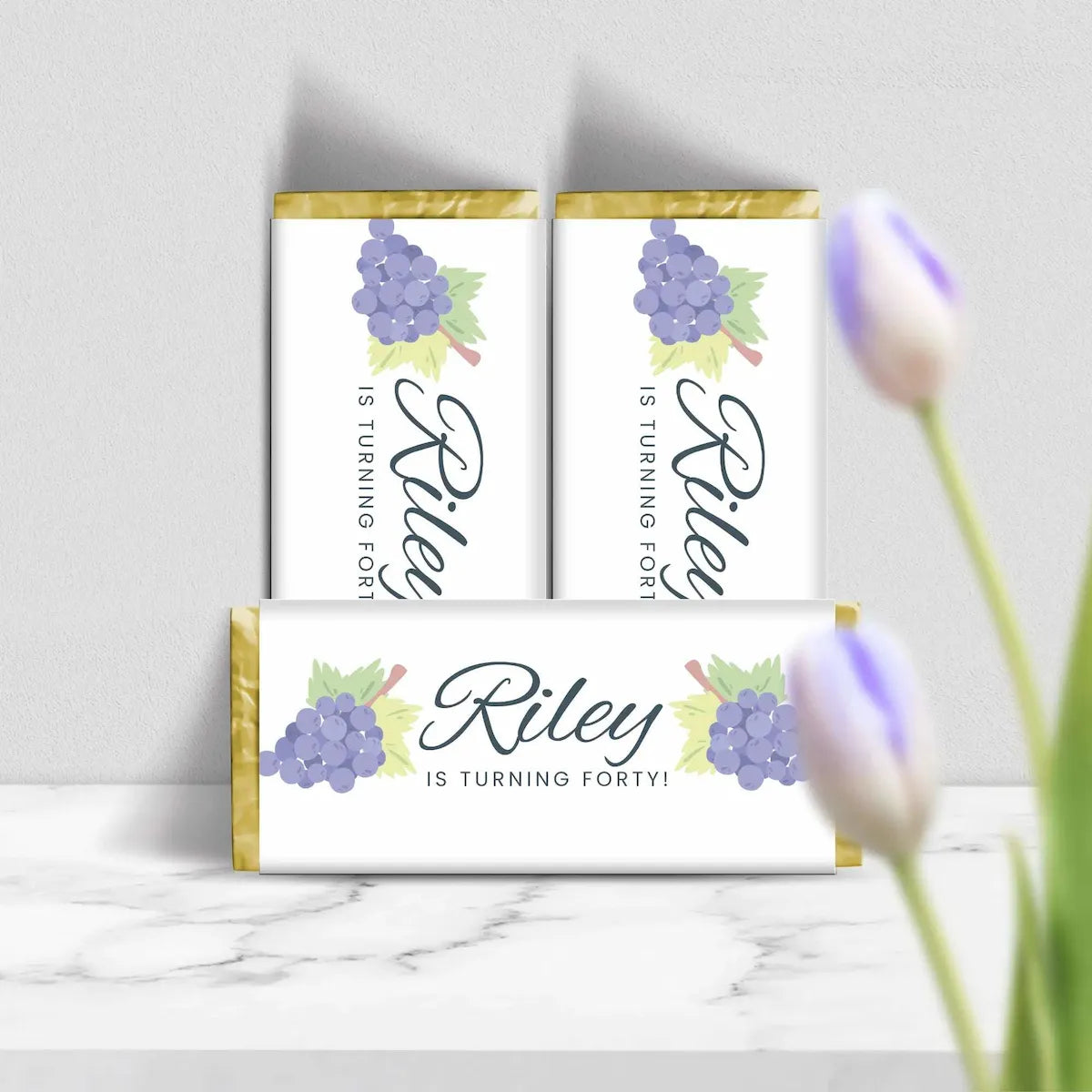 Three personalized chocolate bar wrappers stacked on a marble surface against a light gray wall, with blurred tulips in the foreground. The wrappers are white with gold foil edges, purple grape illustrations, and the text 'Riley IS TURNING FORTY!'.