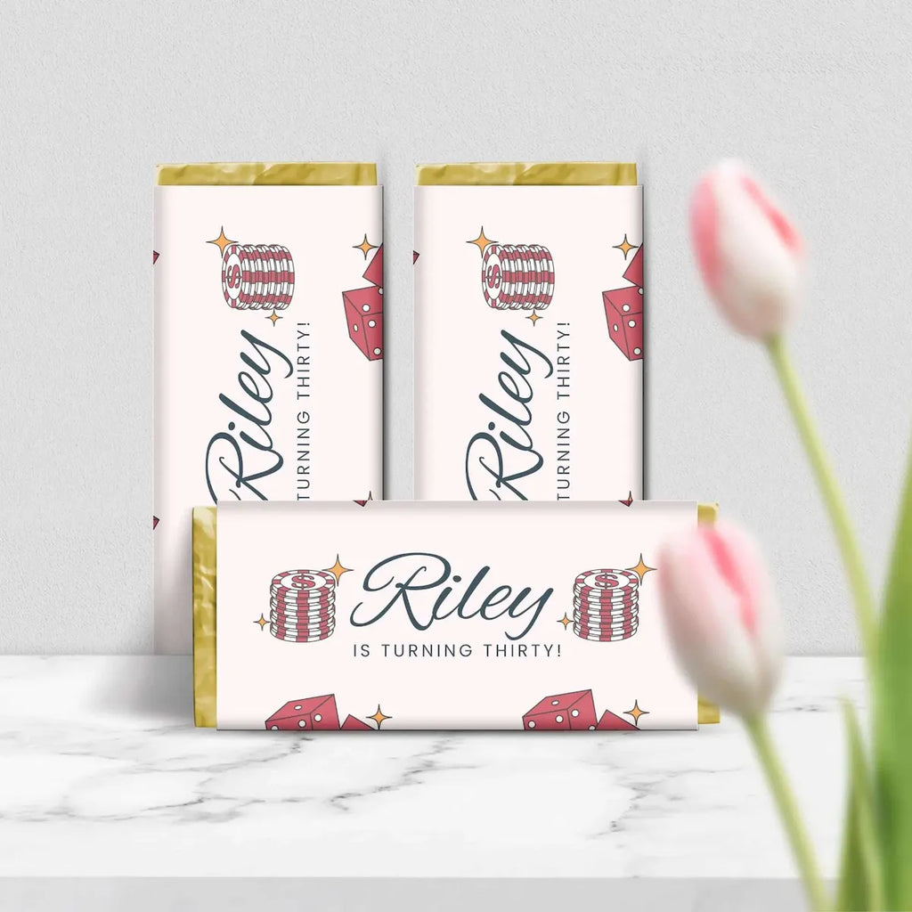 Three personalized chocolate bars with gold foil edges and a white wrapper printed with 'Riley TURNING THIRTY!' and red-and-white striped party hats, displayed on a marble surface with blurred pink tulips in the foreground.