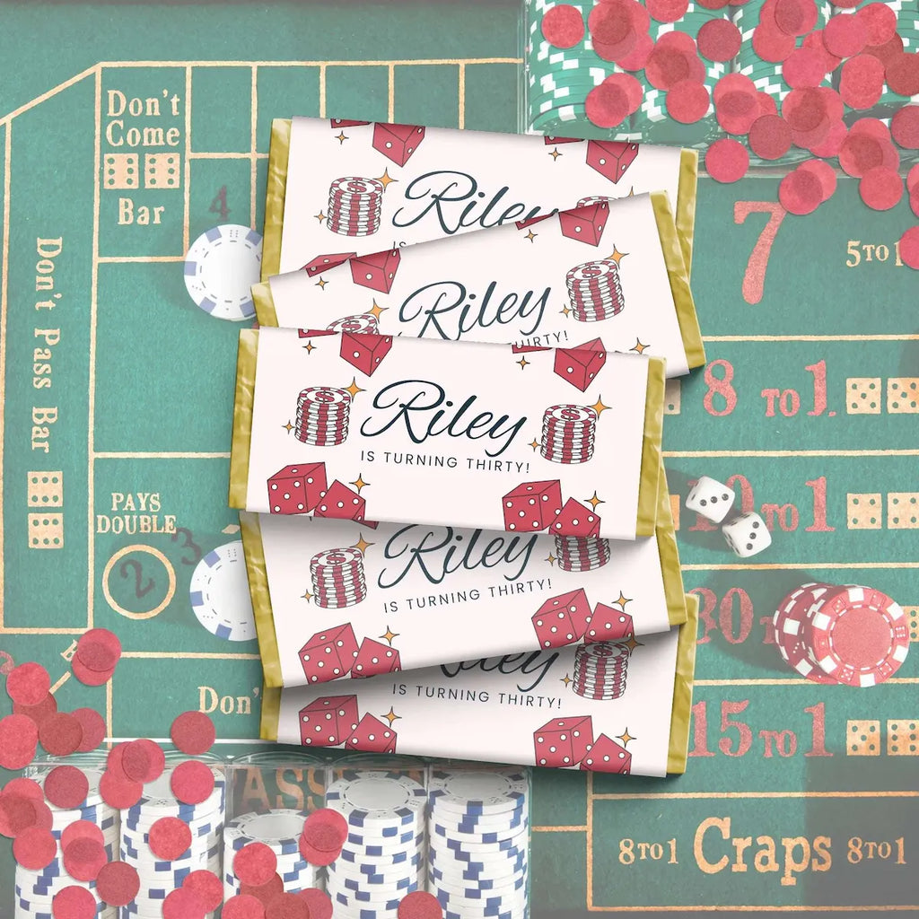 Three personalized chocolate favor bags with a casino-themed design featuring red dice, red chips, and gold borders, laid out on a green craps table with scattered red confetti and dice.