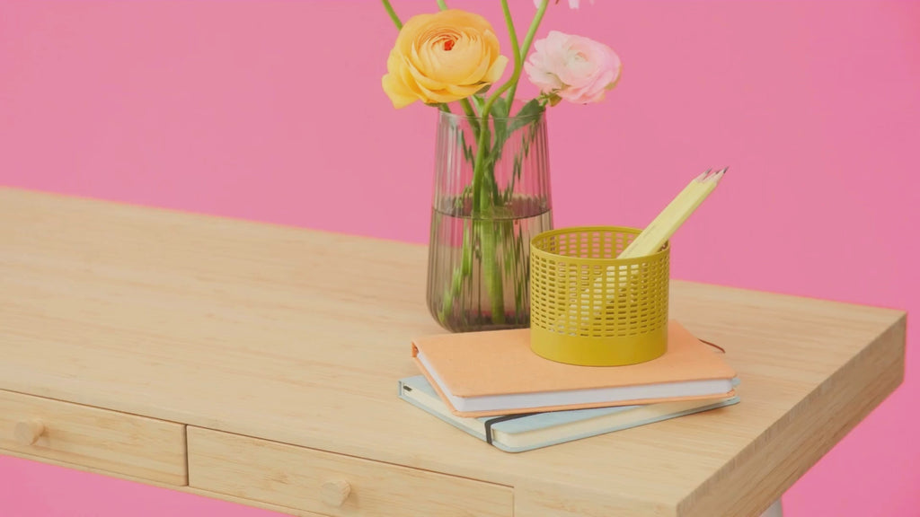 Person putting invite on top of books on wood table with vase of one yellow and one pink flower. 